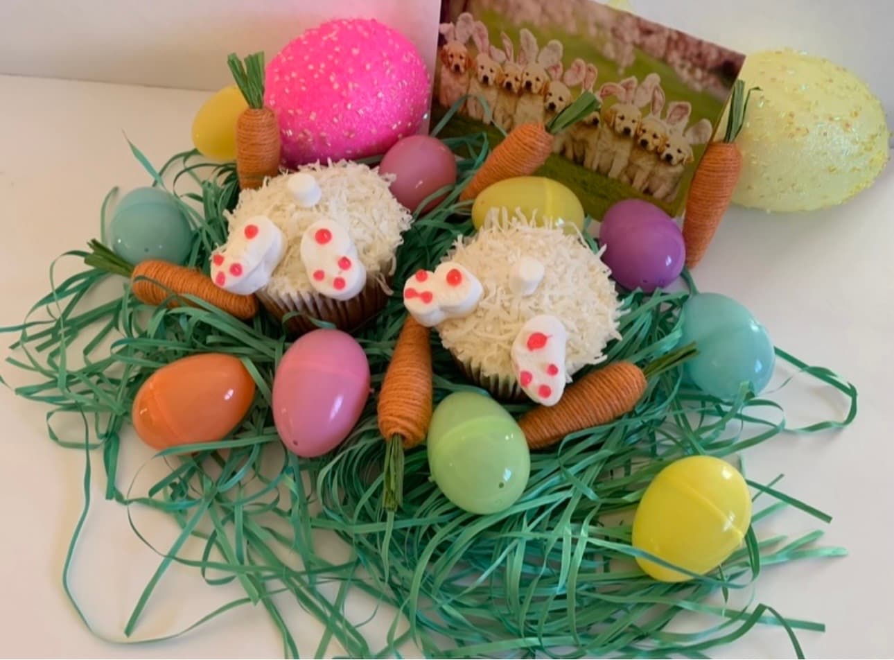 Two bunny-butt cupcakes surrounded by colorful plastic eggs, twine carrots, and green paper grass.