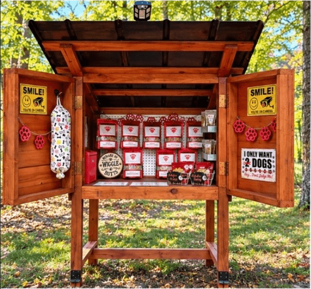 Outdoor wooden stand stocked with bags of dog treats and pet-themed signs.